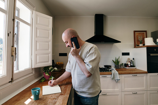Man in kitchen writing in notebook while talking on the cell phone. He is smiling. There is a cup with liquid that may be coffee in front of you. The image is in angle and you can see almost the entire kitchen