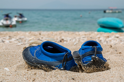 Coral slippers. Summer shoes for stones beach. Blue water shoes. Shoes which protect foot on a beach. Summer vacation background with a pair of sandals on beach. Copy space.
