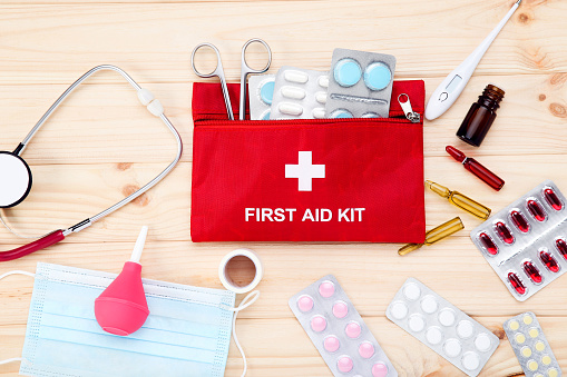 A first aid kit complete with emergency medical supplies sits against a white background.  A red container with a white cross rests in the middle of various first aid supplies including gauze, adhesive bandages, surgical tape, a thermometer, an open container of pills and a stethoscope.