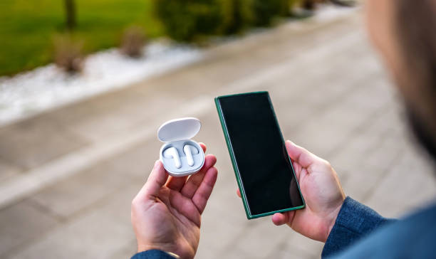 Businessman holding white wireless earbuds in charging box and smartphone outdoors