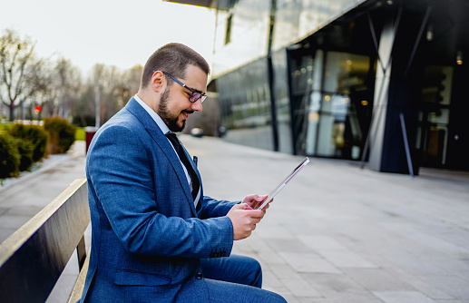 Young cheerful businessman using a tablet while sitting on the bench in front of an office building