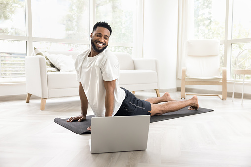 Happy sporty African American guy practicing yoga at laptop, doing upward facing dog pose, backbend asana, looking at screen, smiling, laughing, taking online training class