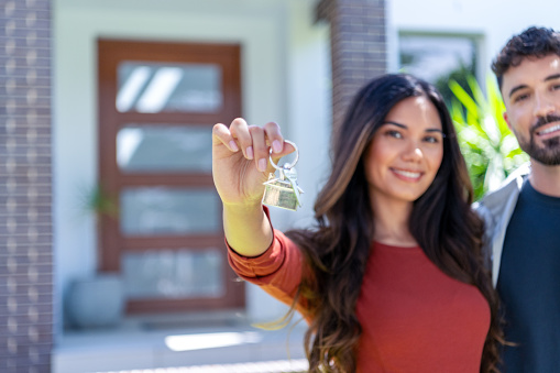 Close up happy woman and man, holding keys from new first house, young family celebrating moving day, satisfied customers couple purchase real estate, mortgage and relocation concept. Front door can be seen in the background Focus on foreground