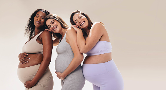 Multicultural pregnant women in fitness clothing attend a prenatal yoga class, leaning on one another for support. Happy and smiling, they prioritize self-care and wellbeing during their pregnancy.