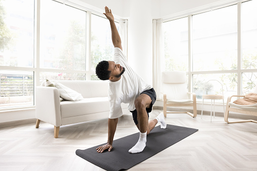Serious young Black man doing morning exercises at home, keeping twisting yoga asana on mat, caring for wellness, fit, wellbeing, healthy active lifestyle, training body in apartment