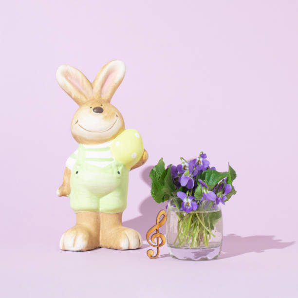 Easter bunny stands next to violets in a vase and a treble clef. Copy space. Pastel purple background. Front view.