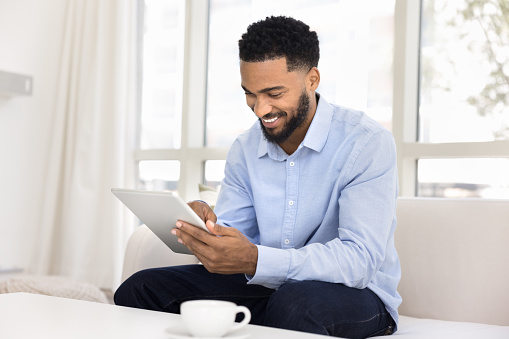 Happy young African entrepreneur man enjoying Internet technology, smart home app on digital tablet, siting on white couch, using online communication for distant work