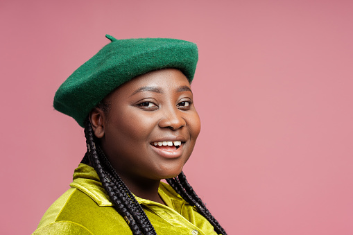 Smiling, happy African American woman with stylish braids hair wearing green beret and casual outfit posing in studio isolated on pink background copy space. Concept of advertisement, shopping