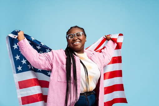 Excited beautiful African American woman, patriot wearing glasses holding American flag isolated on blue background. Attractive female supporter, election day concept