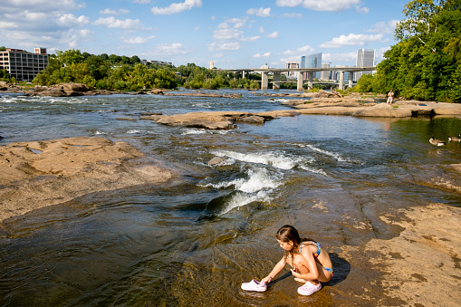 Little girl playing with water on the riverside uses her clogs to refresh herself