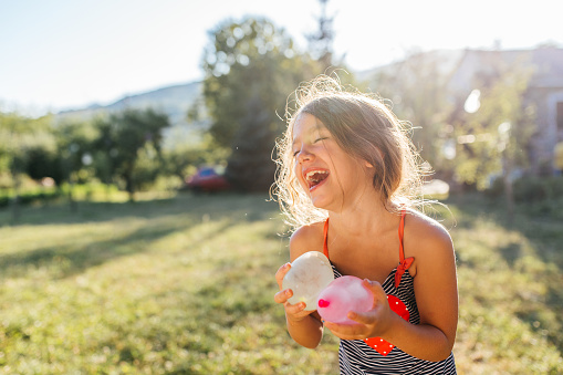 Water balloon games for kids. Smiling girl holding colorful balls filled with water. Cheerful and ready for water fight in the yard on a hot summer sunny day