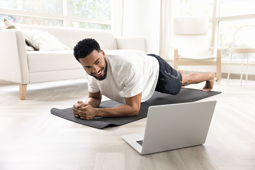 Cheerful strong athletic African man keeping static plank on yoga mat at laptop, looking at screen, smiling, watching lesson, using online communication for fitness workout at home