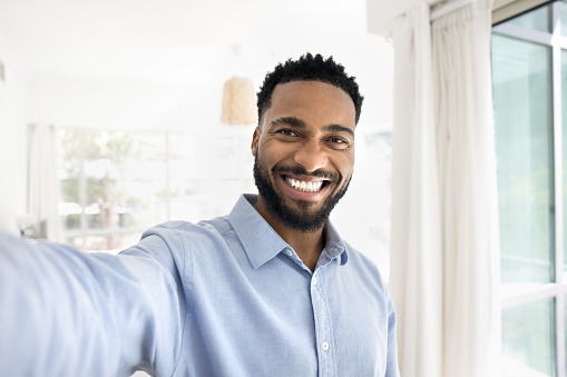 Cheerful handsome young African man taking selfie, looking at camera with toothy smile, showing perfect white teeth, holding gadget with webcam in outstretched hand, talking on video call