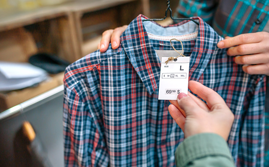 Close up of unrecognizable woman client hand holding label of plaid shirt on clothing shop. Female employee showing tartan shirt on hanger to customer in a store. Selective focus on apparel label.