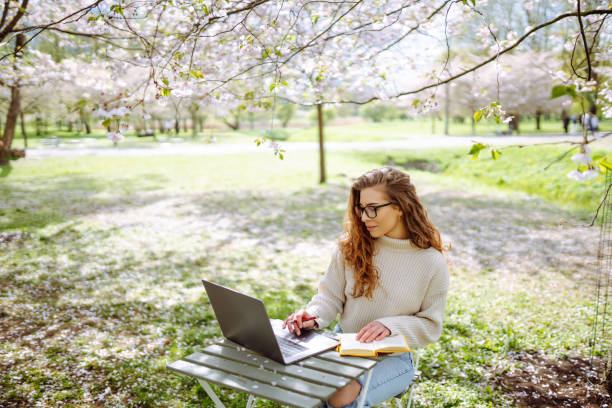 Beautiful woman sitting on blooming park and using laptop.
