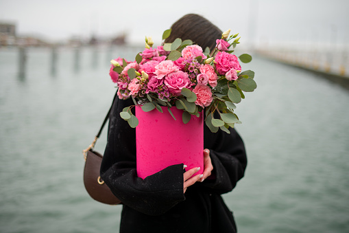 Young woman in a black coat holds a bouquet of flowers in a red gift box on the background of the sea