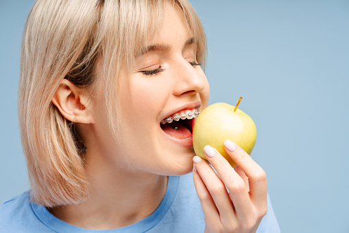 Closeup shot of gorgeous female wearing braces, enjoying an apple, isolated on a blue background. Highlights oral care