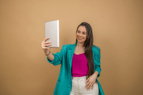 istock Attractive female model with brunette hair is grimacing in studio, making selfies on digital tablet camera on the beige background 2066672389