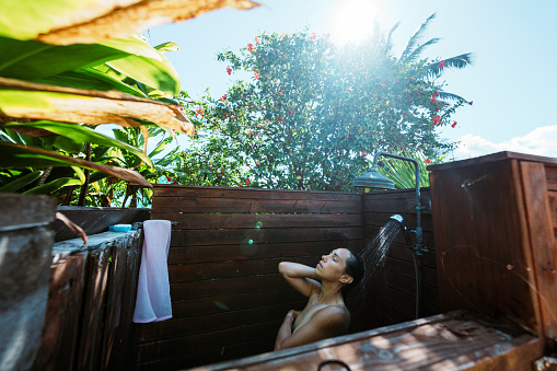 An Eurasian woman of Hawaiian and Chinese descent closes her eyes and rinses conditioner from her hair while taking a relaxing and refreshing shower outdoors in the lush backyard garden of a vacation rental home located in Hawaii.