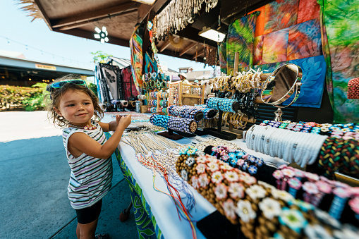 A cute three year old Eurasian girl smiles with excitement while looking at a display of jewelry for sale at a street market in Hawaii.