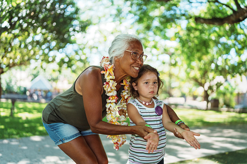 A beautiful and active senior woman of Hawaiian and Chinese descent assists her three year old Eurasian granddaughter as they attend an entry level community hula lesson for all ages located in a public park in Hawaii.