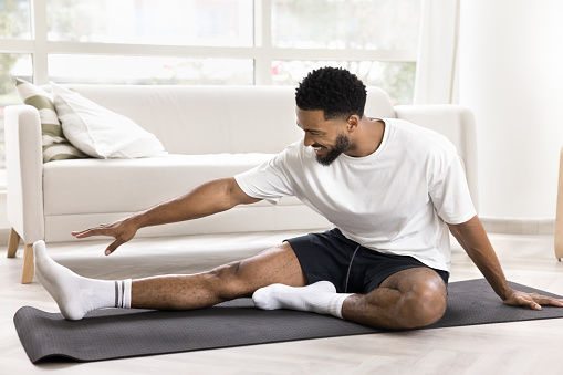 Happy active young Black man keeping fit, training body at home, stretching hip and back on yoga mat, bending forward to leg, smiling, enjoying morning exercises
