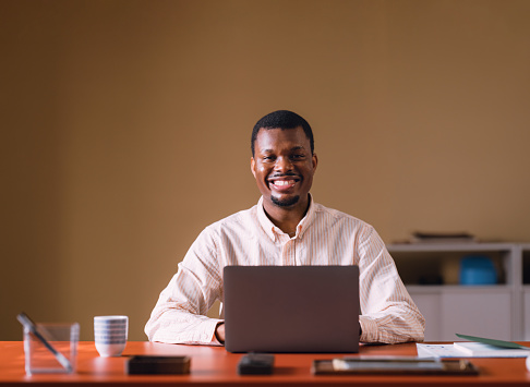 A content professional African man smiling at the camera with a laptop in a well-lit office environment, exuding confidence and positivity.