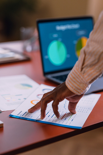 Close-up of a business professional's hands reviewing and analyzing colorful data charts and graphs on documents beside a laptop computer.