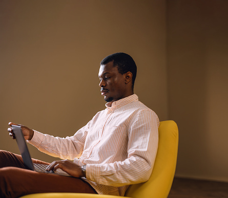 Professional African American male entrepreneur using a computer while seated in a modern yellow chair at home or office.