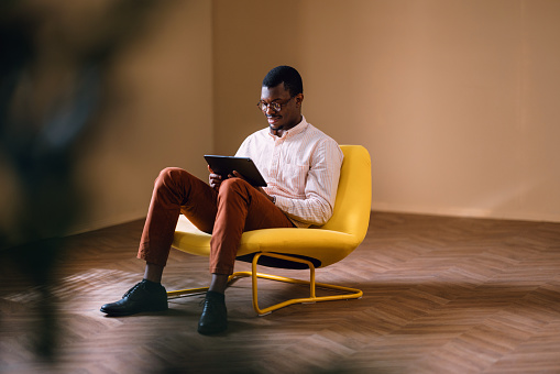 Concentrated young African man sitting in a stylish yellow chair working on his digital tablet in a contemporary room setting.