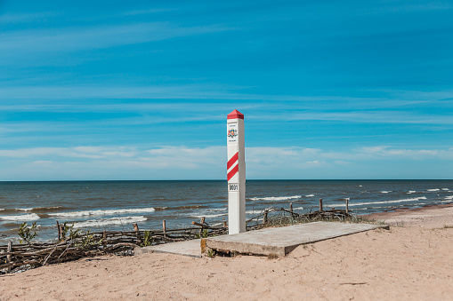 Red and white striped border post marked '0001' on a sunny beach, symbolizing the starting point of the border along the coastline