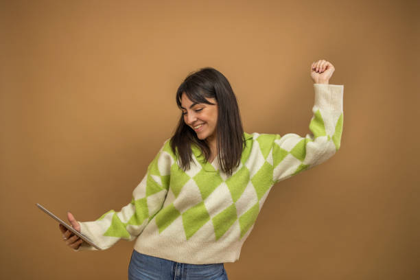 Attractive female model with brunette hair is grimacing in studio, making selfies on digital tablet camera on the beige background