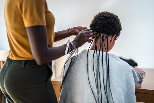 A mixed race female creates braids on a mid adult Black female's hair in a cozy home setting.