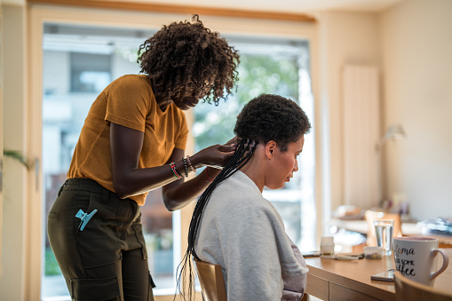 A mixed race female is making braids for a mid adult Black female in a domestic setting.