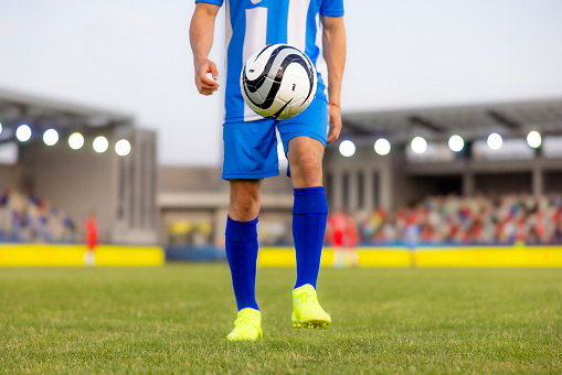 Close-up shot of an unrecognizable male soccer player in blue and white uniform with neon cleats, holding a football while standing on the pitch during an outdoor match.