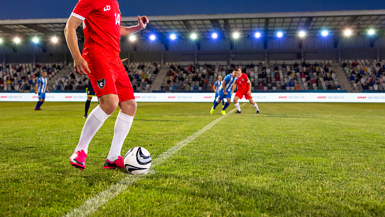 Unrecognizable male soccer player in red gear steadies the ball for kickoff at a professional stadium. The scene captures the anticipation of the game in an outdoor setting at twilight, with the opposing team in the background.