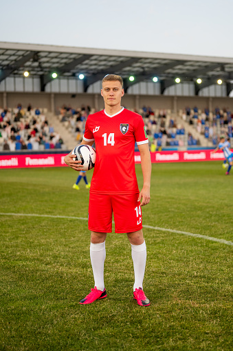 A tall Caucasian male football player in a red uniform holds a soccer ball confidently while standing on the pitch. An outdoor setting with stadium seating and a match in progress is visible in the background. Professional athletic attire and posture.