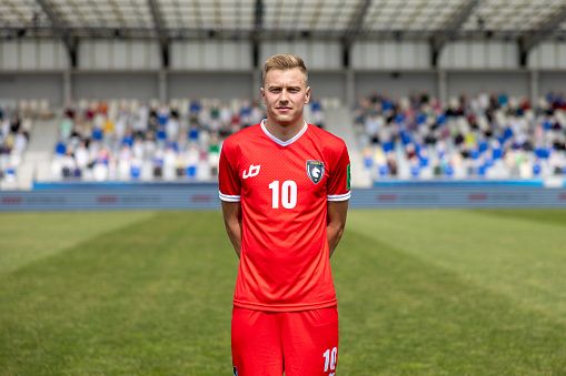 A determined young Caucasian male football player stands in professional red sports attire on the pitch. The stadium seats create a blurred background emphasizing his focused expression. Outdoor, daytime setting.