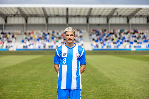 A professional male Caucasian football player stands confidently on the pitch, wearing a blue and white striped uniform with the number 3. The stadium seating in the background is partially filled with spectators.