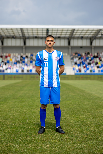 A professional male Hispanic football player stands with a determined look, dressed in a blue and white striped kit. Captured outdoors in a stadium setting, showcasing athletic attire and a competitive spirit.