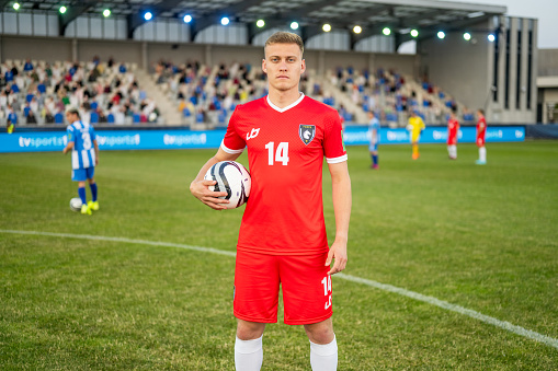 A male Caucasian football player in a red jersey stands holding the ball with determination on the pitch. The stadium setting with spectators and players in the background suggests a professional sports event. His athletic build and focused gaze reflect his readiness for the game.