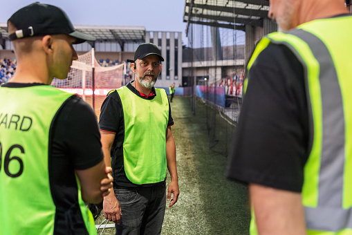 Male security professionals in a high-visibility vest standing guard at an outdoor football stadium during an evening event, maintaining crowd safety and venue security.