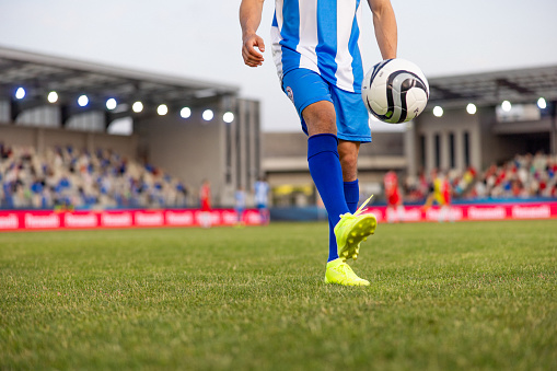Close-up of an unrecognizable male soccer player in professional sportswear dribbling a football on a grass pitch, with stadium and spectators in the background. The scene captures a dynamic outdoor sports event.