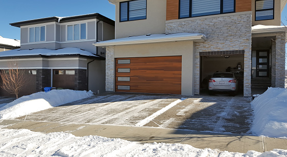 Calgary, Alberta, Canada- March 7,2024:Two newly built homes in winter. Three car front attached garages on both. One open with car inside.  Cleared driveway and walkway at front of home. Stone and concrete siding. Wood paneled garage doors.