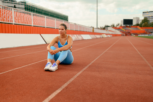 Young beautiful tired caucasian woman in sports outfit sitting and relaxing on a racetrack before or after workout or race competition outdoor in the city stadium. Copy space. Healthy lifestyle.