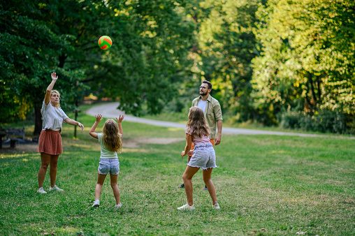 A sportive fun parents playing a game with a ball in nature with their daughters on sunny summer day at weekend. An active family on a meadow near forest is playing a fun game with a ball. Copy space.