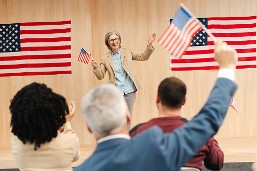 Portrait of smiling confident woman, politician, presidential candidate holding American flag communication, speaking with audience. Vote, United States presidential election concept