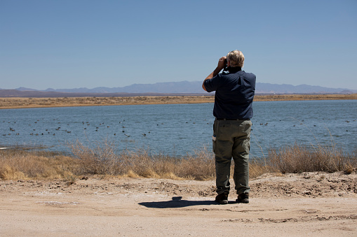 A man with binoculars and a bird book in his pocket is looking at the pond with his binoculars.  A birdwatcher in AZ.
