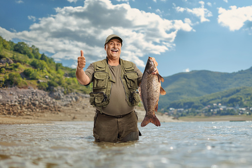 Happy mature fisherman holding a carp fish and gesturing thumbs up inside a lake