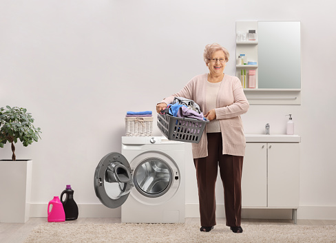 Full length portrait of an elderly lady holding a laundry basket with clothes inside a bathroom